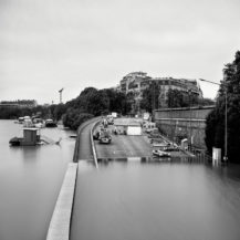 A black and white photo shows a city street partially submerged by floodwater, with cars, boats, and tents along the road. Tall buildings and trees line the background under an overcast sky.
