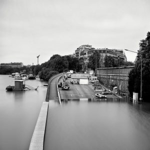 A black and white photo shows a city street partially submerged by floodwater, with cars, boats, and tents along the road. Tall buildings and trees line the background under an overcast sky.