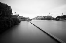 Black and white photo of a calm river with a bridge in the background, buildings and trees on the left, and several construction cranes visible on the right under a cloudy sky.