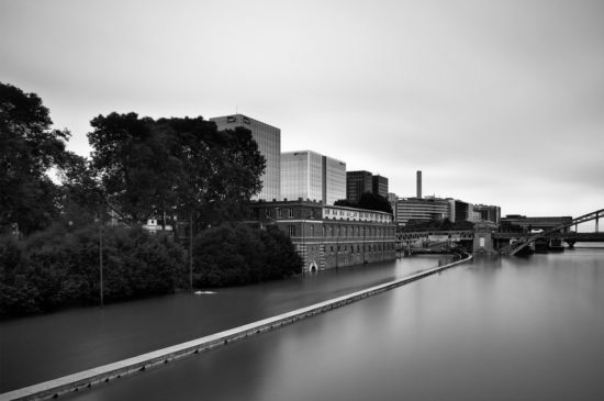 Black and white photo of a riverside cityscape with modern office buildings, older brick structures, trees, and water covering the lower parts of the buildings and walkway, suggesting flooding.