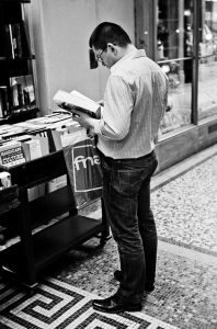 A man stands indoors reading a book beside a table of books. He holds a Fnac shopping bag and wears glasses, a striped shirt, jeans, and loafers. The scene appears to be in galerie Vivienne, perhaps browsing after a lecture.