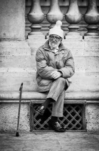 An elderly man wearing a winter coat and a knit hat sits on a stone ledge with his legs crossed, smiling. A cane rests beside him. Decorative balusters and stonework form the background. The image is in black and white.