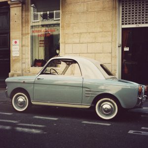A vintage petite voiture in light blue and white is parked on a city street, right in front of a beige stone building with a shop window displaying text and a mannequin inside.