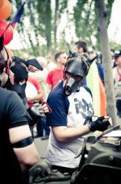 Two people wearing black dog masks and gloves are talking at an outdoor LGBT event in 2016. Other people and trees are visible in the background, with some balloons seen in the upper left corner.