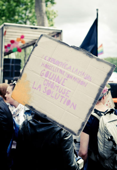 A person at a protest holds a cardboard sign painted with French words in colorful letters. Other people, a truck, and a rainbow flag can be seen in the background, reflecting the vibrant spirit of Gay Pride 2016.