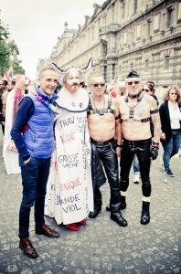 Four people stand together at a Gay Pride 2016 parade, three in elaborate or leather outfits, one in a blue vest and jeans. Old buildings line the street as other Gay Pride participants are visible in the background.