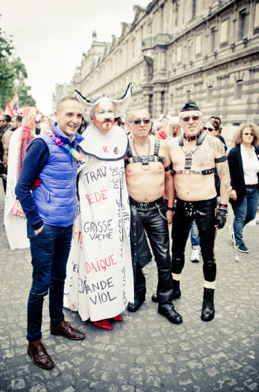 Four people stand together at a Gay Pride 2016 parade, three in elaborate or leather outfits, one in a blue vest and jeans. Old buildings line the street as other Gay Pride participants are visible in the background.