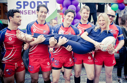 Five smiling rugby players in red uniforms hold up a woman in a blue outfit, who is laughing and holding a rugby ball. Colorful balloons and a "SPORT" sign create a festive Gay Pride Parade atmosphere in the background.