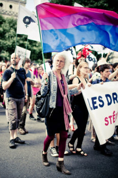 An older person with white hair holds a large bisexual pride flag while marching in a Gay Pride Parade. Other people hold signs and banners in the background, and the atmosphere is energetic and joyful.