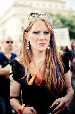 A woman with long blond hair, wearing a black top, butterfly necklace, and earrings stands in a crowd at an outdoor Gay Pride 2016 event, looking focused. People and a blurred sign are visible in the background.