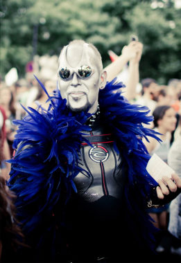 A person in a metallic costume with reflective glasses, white face paint, and a bold blue feather boa stands among a crowd at an outdoor event, holding a piece of paper during Gay Pride 2016.