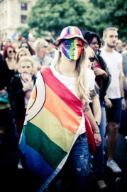 A person wearing a rainbow-striped hat, a rainbow flag draped over their shoulders, and a mask painted with the LGBTQ+ pride colors stands among a crowd at an outdoor Gay Pride 2016 event.