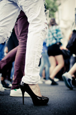 A person wearing ripped white jeans and black high heels walks on a street during Gay Pride 2016, surrounded by others in casual clothing, some blurred in the background.
