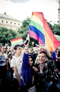 A group of young people celebrate at Gay Pride 2016, smiling and holding rainbow flags. One person holds up a phone, while others make peace signs. The background shows more participants and a large building.