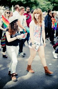 Two young women walk in a crowded Pride parade; one wears a hat and reads a flyer, the other wears a t-shirt, shorts, and boots, holding a bag. People around them carry rainbow flags at Gay Pride 2016.