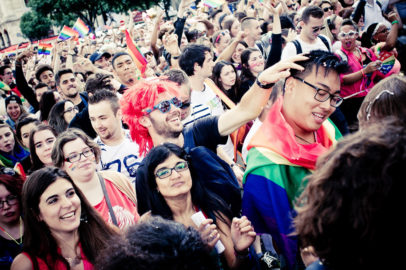 A large, joyful crowd at a Pride Parade, with people smiling, wearing colorful clothing, waving rainbow flags, and celebrating Gay Pride together outdoors.