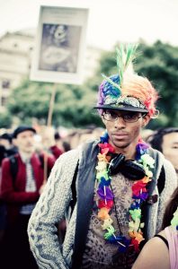 A person wearing a rainbow-colored wig, glasses, headphones, a knit sweater, and a colorful flower lei stands in a crowd at an outdoor Gay Pride event. A blurry sign is visible in the background.