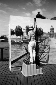 A person in light clothing and a hat stands on a small platform, painting a realistic outdoor scene with an expert coup de pinceau on a large canvas that blends seamlessly into the actual landscape behind it.