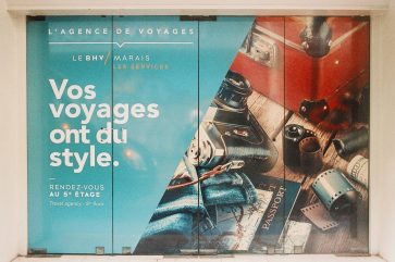 Travel agency ad with a blue background, showcasing a passport, camera, and map. White French text says, "Vos voyages ont du style," inspiring stylish voyages, plus agency details and location info.