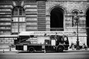 A black and white photo of a camion de pompier parked before an ornate, historic stone building in Paris, with a statue in an archway and scooters nearby. Orange cones line the street in front of the truck.