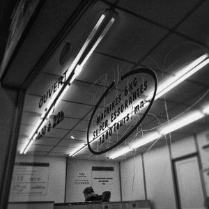 Black and white photo of a Laverie de nuit window with handwritten signs in French advertising 6kg machines, fast spin cycles, and the price “1,20 €”. Reflections reveal ceiling lights and a person inside.