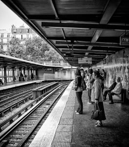 People wait on a covered train platform at Métro Bastille, next to railway tracks. Some stand, some sit on benches, and buildings are visible in the background. The image is in black and white.