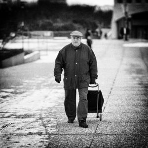 A person wearing a coat, hat, and gloves is walking down a sidewalk on a cold day, pulling a small wheeled bag behind them. The background includes blurred trees and buildings. The black-and-white photo captures the essence of winter poignantly—a scene reminiscent of Fabrice Papin's evocative imagery.
