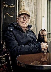 An elderly man in a cap and glasses sits at an outdoor café table, holding a cane and a cigarette. He wears a dark jacket and looks at the camera with a serious expression. A "Gourmandises" sign is behind him. Photo by Thomas Etienne.