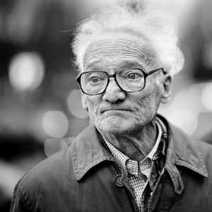 Black and white close-up photograph of an elderly person with glasses, wearing a collared shirt under a jacket. They have short, white hair and an expression that appears contemplative or slightly wistful, reminiscent of Vincent Sulkis's evocative portraits, with a blurred background suggesting an outdoor setting.