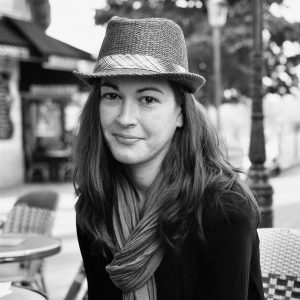 A young woman with long hair wearing a fedora hat and scarf smiles softly while sitting at an outdoor café. The image is in black and white, with a blurred street and café seating in the background.