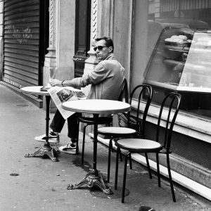 Boris Ballazeau, wearing sunglasses, sits alone at a small sidewalk café table, reading a newspaper. Empty chairs and tables surround him, with a bakery window full of pastries behind him on the quiet, empty street.