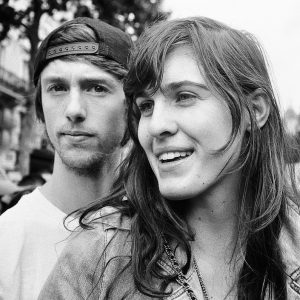 A black-and-white photo of Roman in a backwards cap and Emanuelle with long hair, both looking in different directions. Emanuelle is smiling, while Roman has a neutral expression.