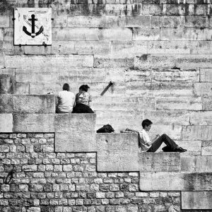 Black and white image of three people sitting sur les marches built into a brick wall. Two people sit together, facing away from the camera, while one sits alone, looking sideways at a device. An anchor symbol is visible on the wall above them.