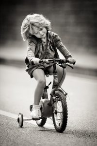 A petite fille with short, tousled hair smiles while riding a tricycle on a paved road. The black-and-white image captures a joyful and carefree moment.