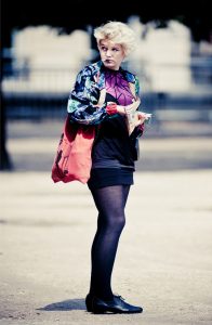 Muriel Nallet, with short blond hair, stands outdoors holding papers, wearing a patterned jacket, purple top, black skirt and tights, black shoes, and a large coral tote bag slung over one shoulder.