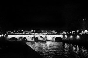 The Pont-Neuf arches are illuminated at nuit, their lights shimmering on the dark river below, with surrounding buildings faintly visible in the background.