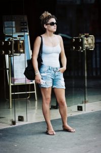 A woman wearing sunglasses, a white tank top, denim shorts, and sandals stands outside glass doors, listening to earphones and holding her phone—her style reminiscent of Natacha Valencay—with one hand in her pocket and a black bag over her shoulder.