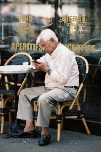 An older man with white hair sits alone at an outdoor café table, reading or using a mobile device. He wears a white shirt and beige pants. Behind him, glass windows display gold lettering advertising wines and beers.