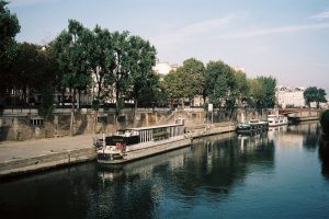 Boats are moored along the edge of the calm Seine River, with trees lining the opposite bank. Buildings and a bridge are visible in the background under a clear blue sky near Quai de Montebello in Paris.