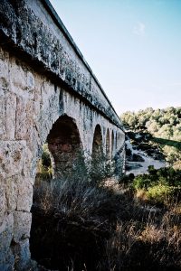 The Aqueduc de las Ferreres, also known as the pont du Diable, is a stone aqueduct with multiple arches stretching across a dry, grassy landscape near Tarragone under a clear, blue sky.