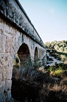 The Aqueduc de las Ferreres, also known as the pont du Diable, is a stone aqueduct with multiple arches stretching across a dry, grassy landscape near Tarragone under a clear, blue sky.