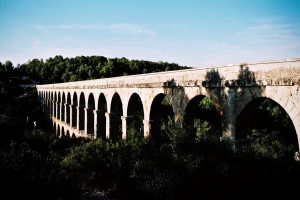 An ancient stone aqueduct, known as the Aqueduc de las Ferreres or pont du Diable, with multiple arches stretches across a green, tree-covered landscape near Tarragone under a blue sky. Shadows from the arches fall on the surrounding vegetation.