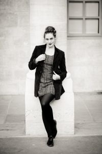 Clotilde, dressed in a plaid dress, dark blazer, and tights, leans against a stone column at the Jardin du Palais-Royal. Captured in noir et blanc, she gazes at the camera with a slight smile, one leg crossed over the other.