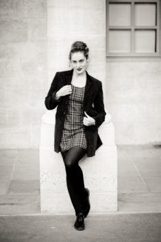 Clotilde, dressed in a plaid dress, dark blazer, and tights, leans against a stone column at the Jardin du Palais-Royal. Captured in noir et blanc, she gazes at the camera with a slight smile, one leg crossed over the other.