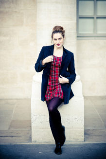A woman with her hair in a bun leans against a stone pillar at Jardin du Palais-Royal, wearing a red plaid dress by Couleurs, a dark blazer, black tights, and black shoes. Pale stone walls and a window frame Clotilde’s Parisian look.