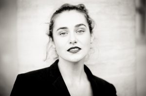 Black-and-white portrait of Clotilde with dark hair tied back, expressive light-colored eyes, and dark lipstick. She is wearing a dark blazer and gazes directly at the camera with a neutral expression, standing against a light-colored background reminiscent of Jardin du Palais-Royal.