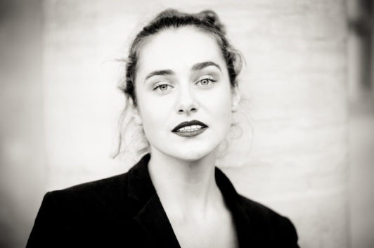 Black-and-white portrait of Clotilde with dark hair tied back, expressive light-colored eyes, and dark lipstick. She is wearing a dark blazer and gazes directly at the camera with a neutral expression, standing against a light-colored background reminiscent of Jardin du Palais-Royal.