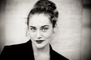 Noir et Blanc portrait of Clotilde with light eyes, dark lipstick, and hair tied up, wearing a dark jacket and gazing confidently at the camera against a blurred Jardin du Palais-Royal backdrop.