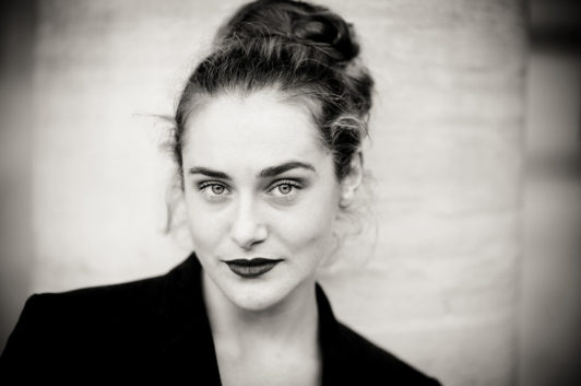 Noir et Blanc portrait of Clotilde with light eyes, dark lipstick, and hair tied up, wearing a dark jacket and gazing confidently at the camera against a blurred Jardin du Palais-Royal backdrop.