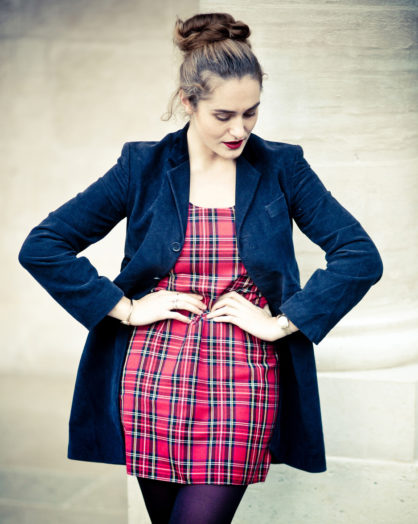 A woman stands confidently, looking down with her hands on her waist. Clotilde wears a red plaid dress with a blue blazer, dark tights, and her hair in a bun. The light, minimalist background evokes the elegance of Jardin du Palais-Royal.